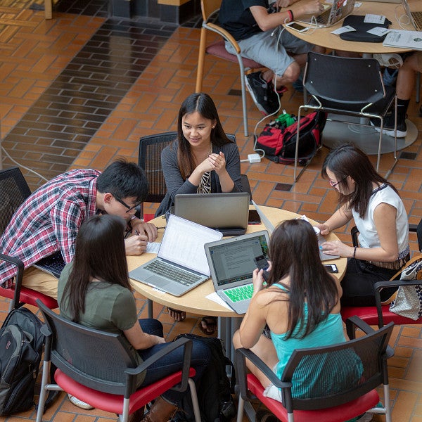 Five students sitting around a table with laptops in the Lillis Business Complex atrium smiling and working on projects.