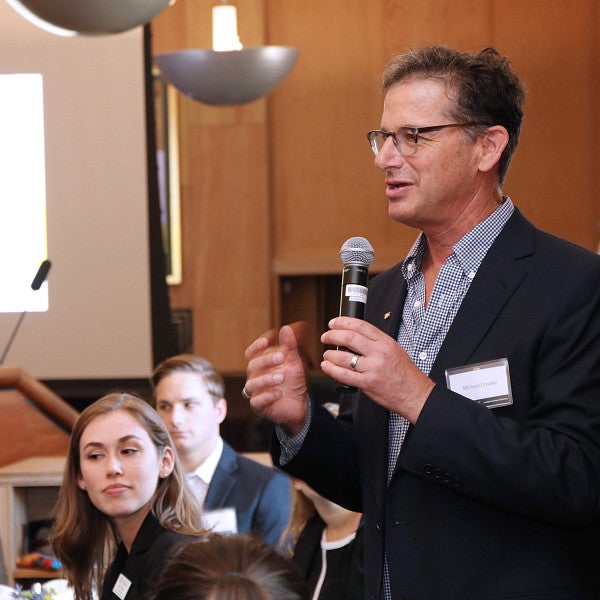 Professor standing and talking in a microphone to students seated at tables.