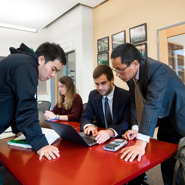 Three students and one faculty member standing around and sitting at a table, looking at a laptop.