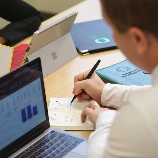 Students sitting at a table with a laptop, writing on a notepad