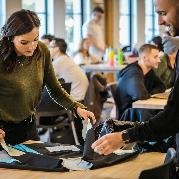 Two students in the foreground working with material at a table with several other students in the background sitting at tables and working on projects