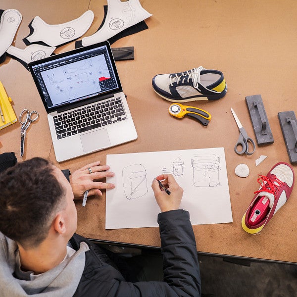 Overhead view of a student drawing on paper surrounded by a laptop, supplies, materials, and athletic shoes