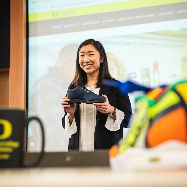 Student at front of the class standing in front of a large screen holding up an athletic shoe