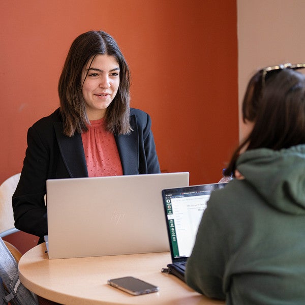 Two students sitting at a round table talking and working on laptops