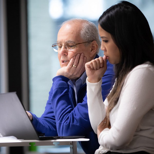 A professor and student standing at a table looking at a laptop together