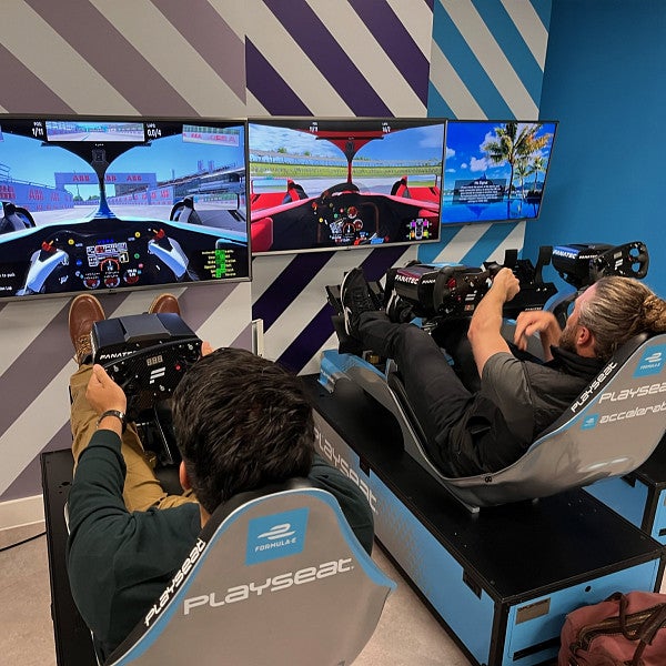 Two MBA students are seated in arcade-style racing seats in front of wall-mounted monitors which display the driver's interior view of a car.