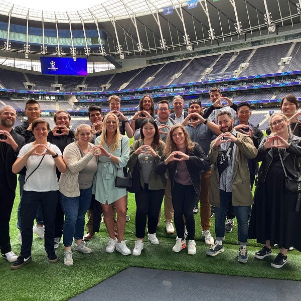 A group of MBA students pose for a photo in a stadium, making the University of Oregon "O" symbol with their hands. They are standing on the stadium turf. The empty stands and stadium awning can be seen behind them.