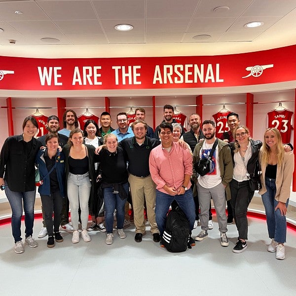A group of MBA students pose for a photo together in a circular room with bright red accents. At the top of the room the words "We are the Aresenal" are painted. Behind the students, several red football jerseys are displayed with different players names