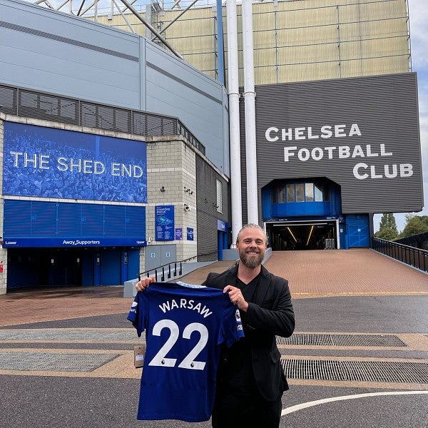 Warsaw Sports Marketing Center program manager Robert Greisinger smiles and holds up a blue number 22 jersey with the word "Warsaw" in place of a player's name. Behind Greisinger is a large building; on the right side the words "Chelsea Football Club" are
