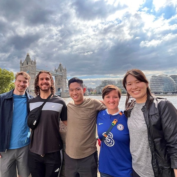 A group of five MBA students smile and pose for a photo in front of the river Thames. Behind them are the Tower Bridge and London's Old City Hall.