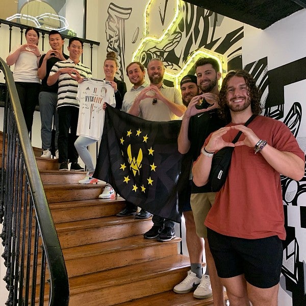 Eight MBA students and a staff member are standing along a staircase from bottom to top, making the University of Oregon "O" symbol with their hands. Several of them are holding a up black flag with a yellow sigil. Another student is holding up a white shirt with text on it.