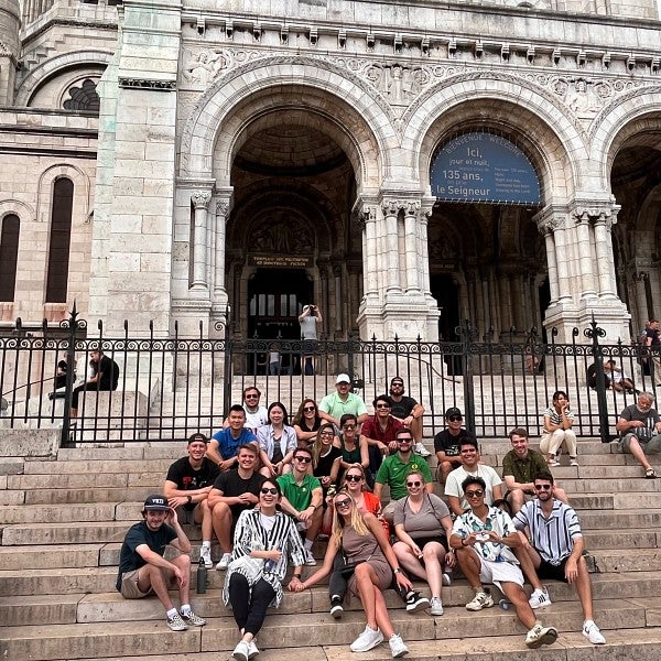 A group of MBA students are seated on the steps of a cathedral in Paris. Behind them, between the steps and the cathedral entrance, is a black iron fence. Statues and other designs can be seen on the exterior of the cathedral.