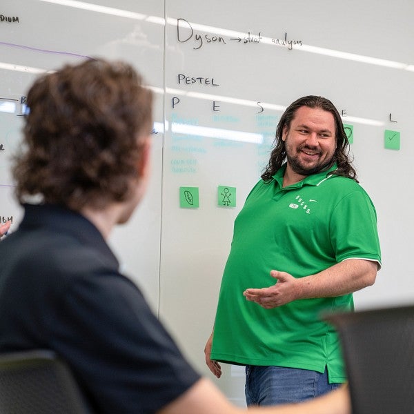 One student wearing a green shirt standing at a white board, facing a classroom and one student sitting, looking at the whiteboard