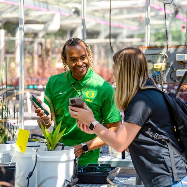 Two students in a greenhouse, studying plants and looking at each others phones while smiling