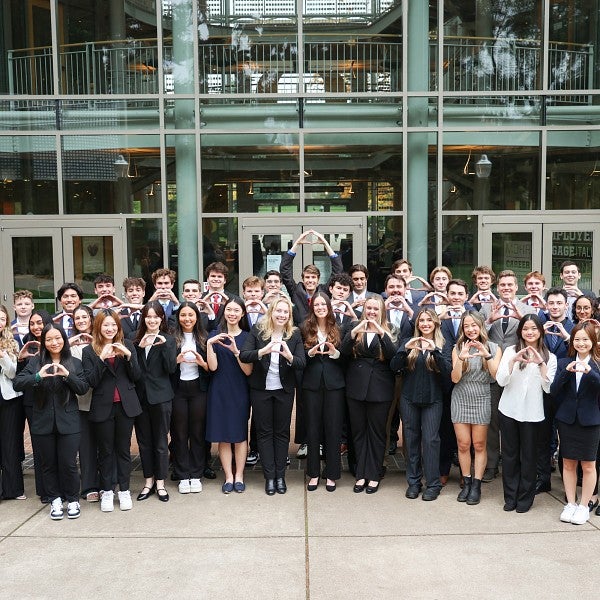 A group photo of business honors students standing in front of the Lillis Business Complex holding their hands in the shape of an O.