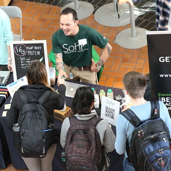 Several students gathered around an alum, talking and shaking hands, tabling at an event,