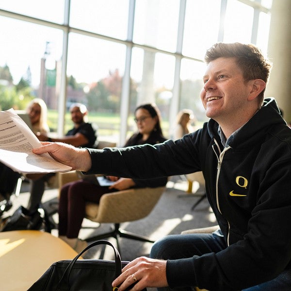 A student sitting in the foreground smiling and holding a small stack of papers. Several students sit in the background in soft seating with a large wall of windows behind them.