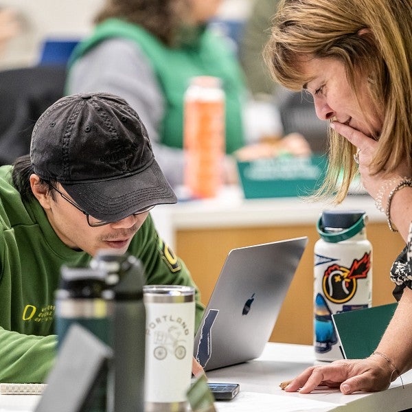 One student sitting down at a desk looking at a computer while another student is standing on the other side of the desk leaning towards the first student.