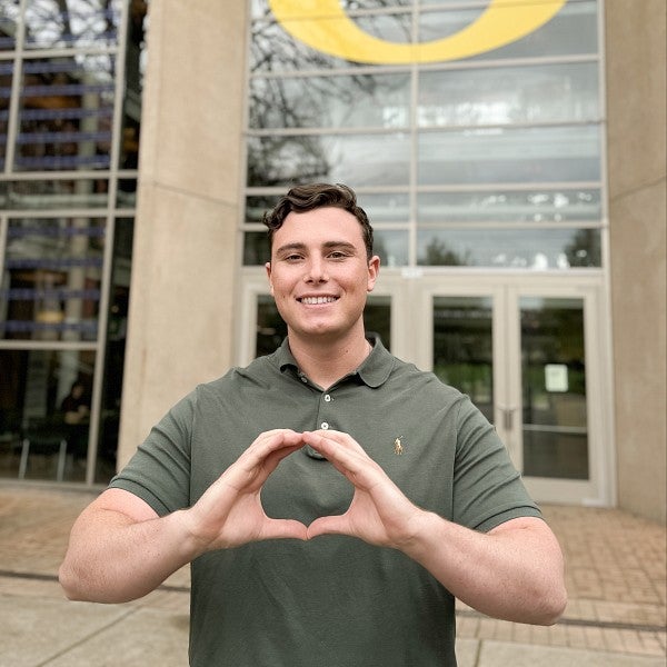 Alex La China standing in front of the Lillis Business Complex smiling and throwing the O.