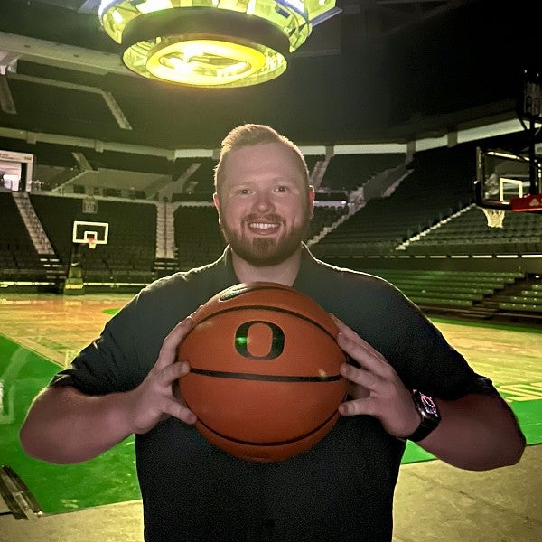 Photo of Quinn Van Horne, standing courtside inside Matthew Knight Arena, smiling at the camera and holding a UO-branded basketball.