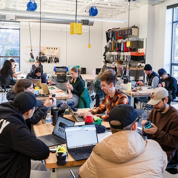 A room full of students sitting around work tables with laptops talking and working on projects