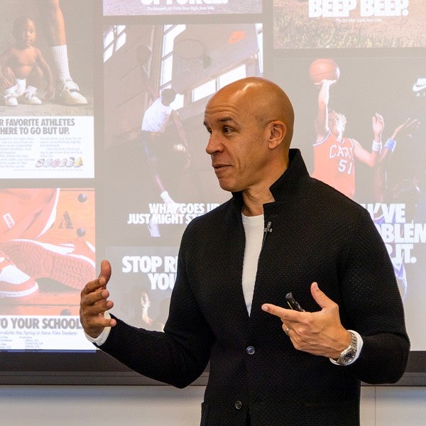 A professor standing at the front of a classroom talking and gesturing with his hands. There is a screen behind him showing several examples of sports industry ads.