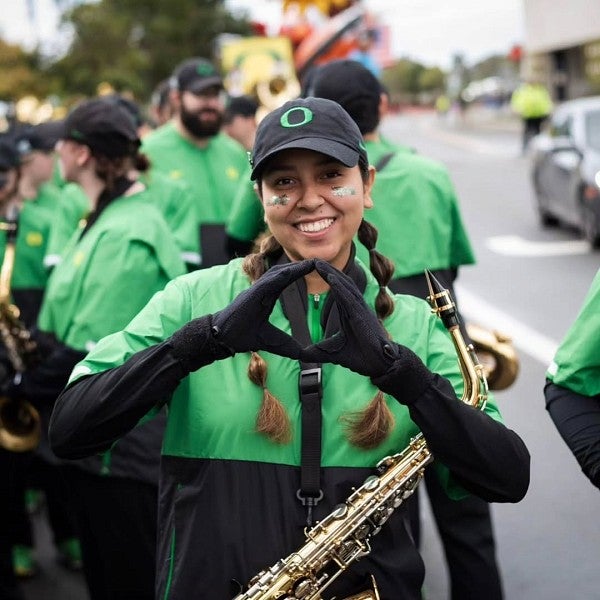 Victoria Castillo is standing outside in a crowd dressed in Duck gear and holding a saxophone while smiling at the camera and throwing the O in excitement.