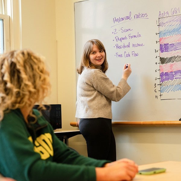 A close-up of a student standing at and facing a whiteboard with a pen in their hand gesturing at accounting figures. Their their head is turned back towards two students sitting at a table. 
