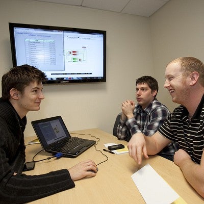 Three students sit at a small table around a laptop with a dispplay screen in the background while assessing a product