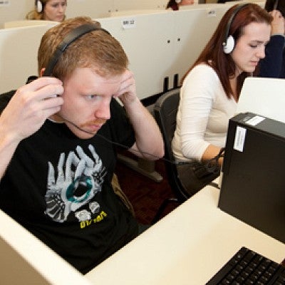 A student sitting at a computer cubicle leans forward while putting on a headset in preparation for a study